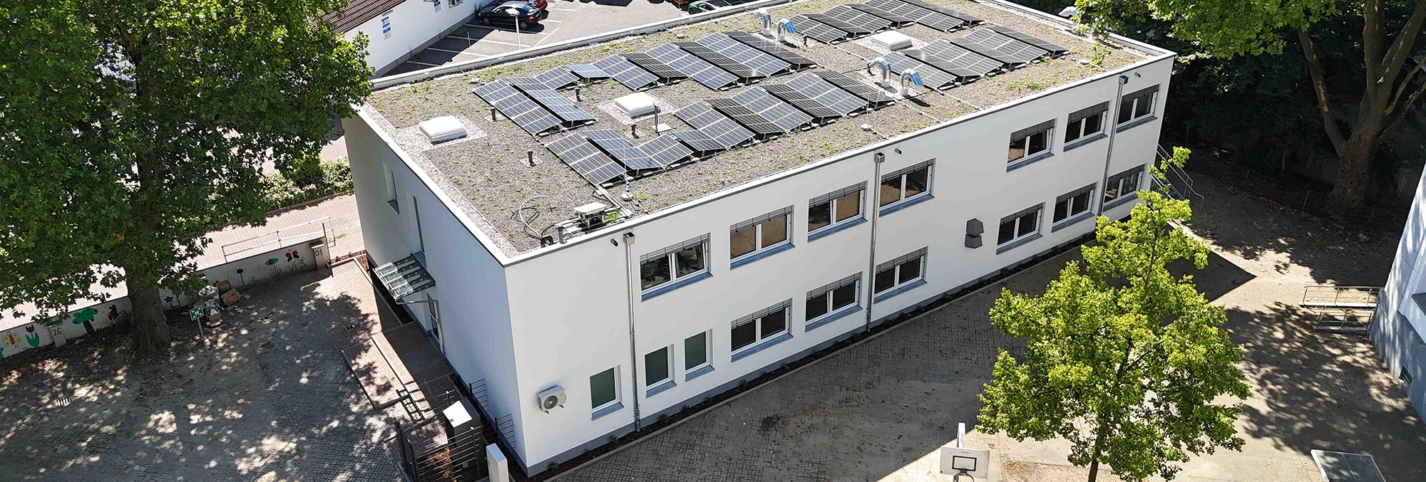 Zweistöckiges weißes Gebäude trägt mehrere Solarpaneele auf dem Flachdach und steht in einem gepflasterten Innenhof, umgeben von Bäumen, Schattenflächen und einigen parkenden Autos im Hintergrund.