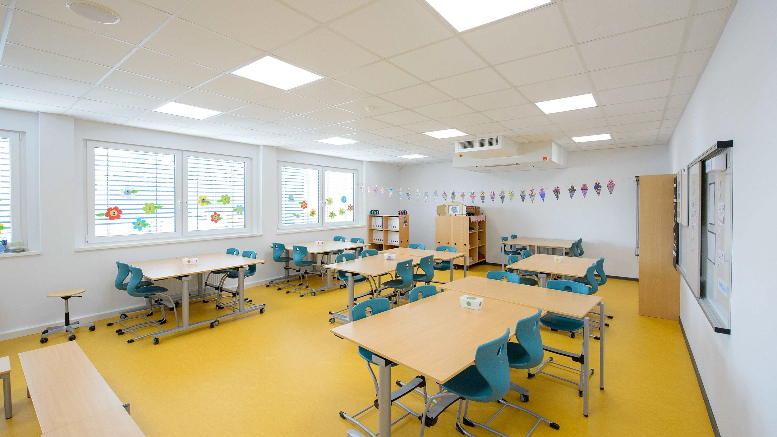 Tische und türkisfarbene Stühle stehen geordnet in Gruppen, bereit zum Unterricht; Fenster mit Blumenaufklebern, gelber Boden, Bücherregale und Girlanden an den Wänden in einem hellen, modernen Klassenraum.