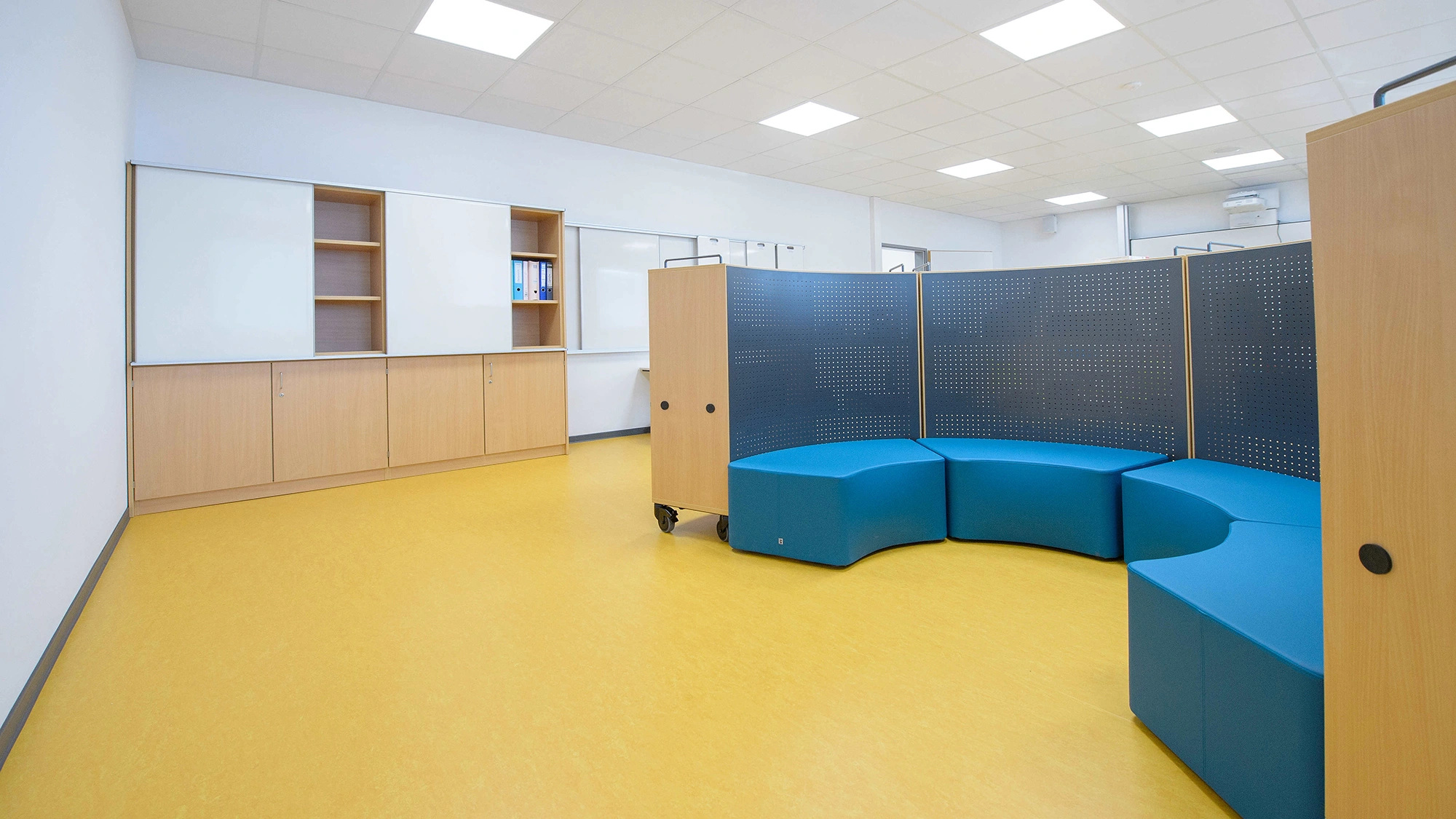 Blaue modulare Sitzmöbel bilden eine halbrunde Sitzecke; sie stehen vor einer perforierten Trennwand in einem hellen Raum mit gelbem Boden, weißen Wänden und niedrigen Holzschränken.
