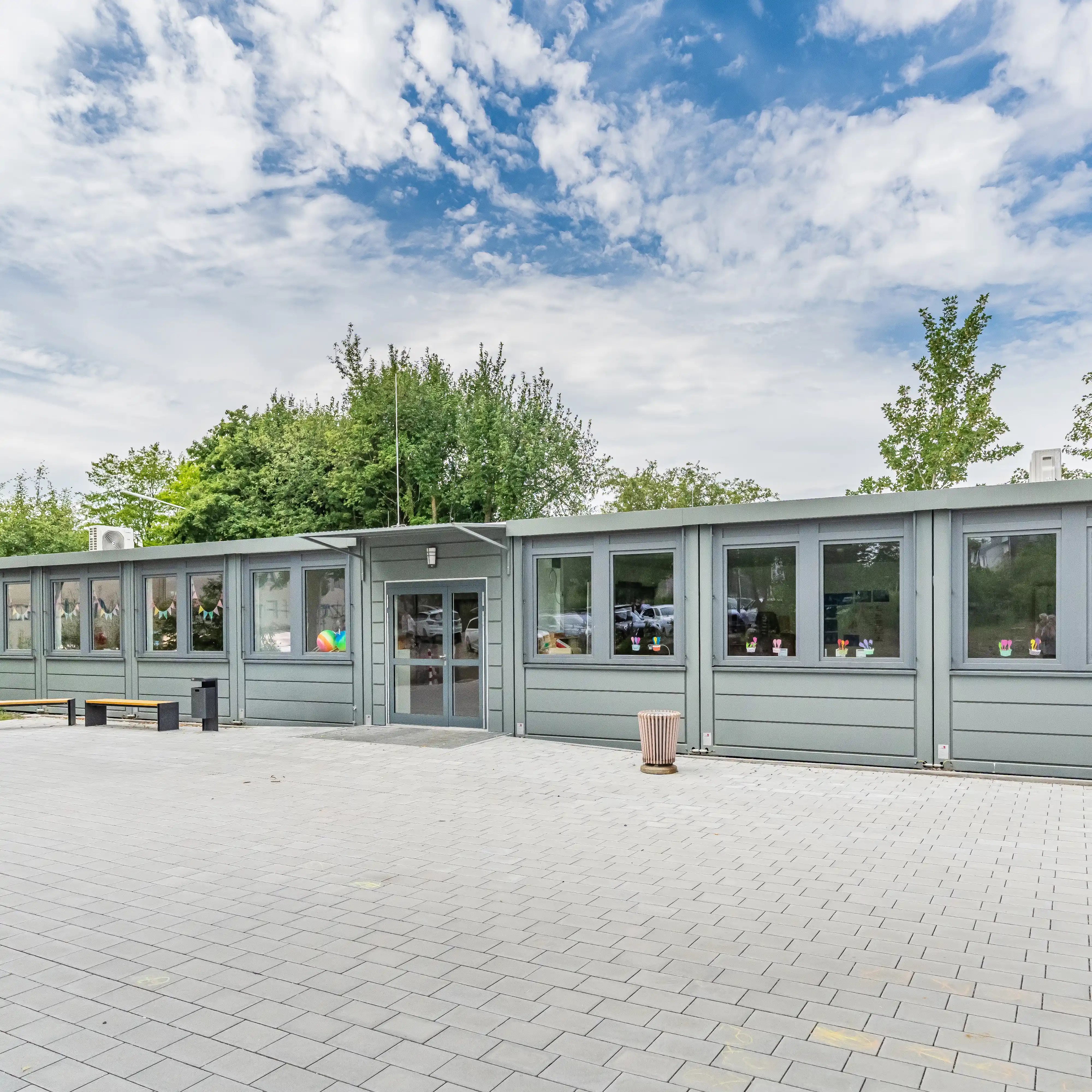 Weitläufiger gepflasterter Vorplatz vor der Containeranlage des Förderzentrums Vohenstrauß mit grauer Fassade, Sitzbänken und Bäumen im Hintergrund