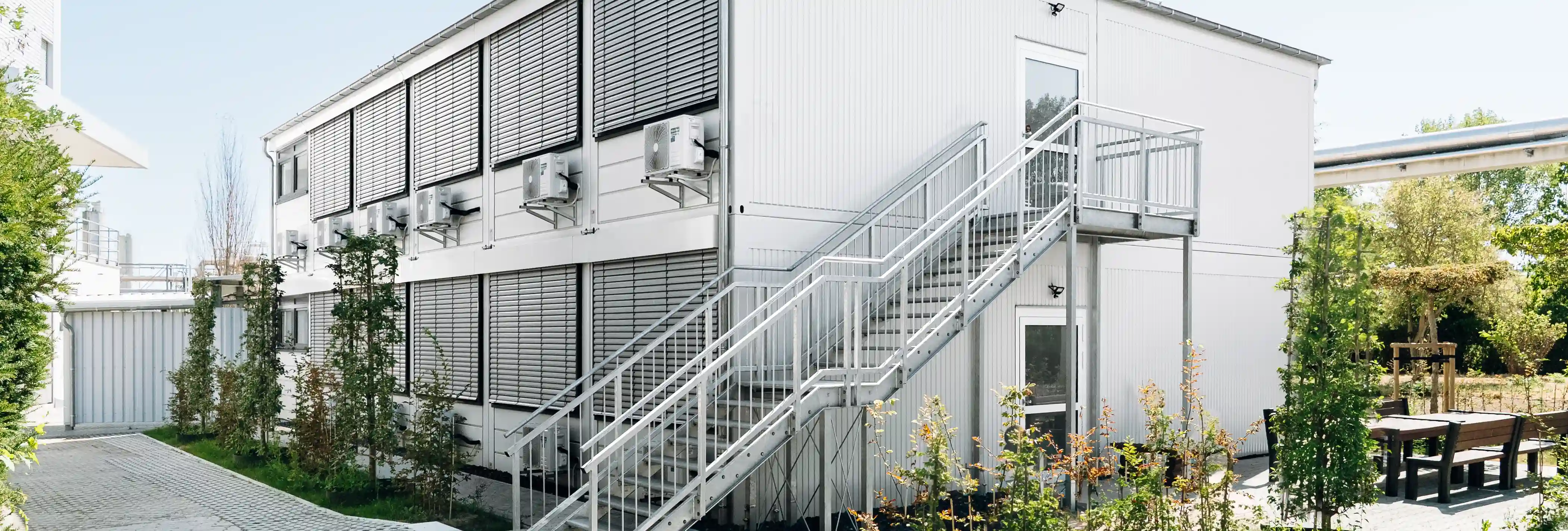 Zweigeschossiges Containergebäude mit außenliegender Treppenanlage, Sonnenschutzsystemen und moderner Gebäudetechnik. Das Gebäude ist von Grünflächen und befestigten Wegen umgeben.