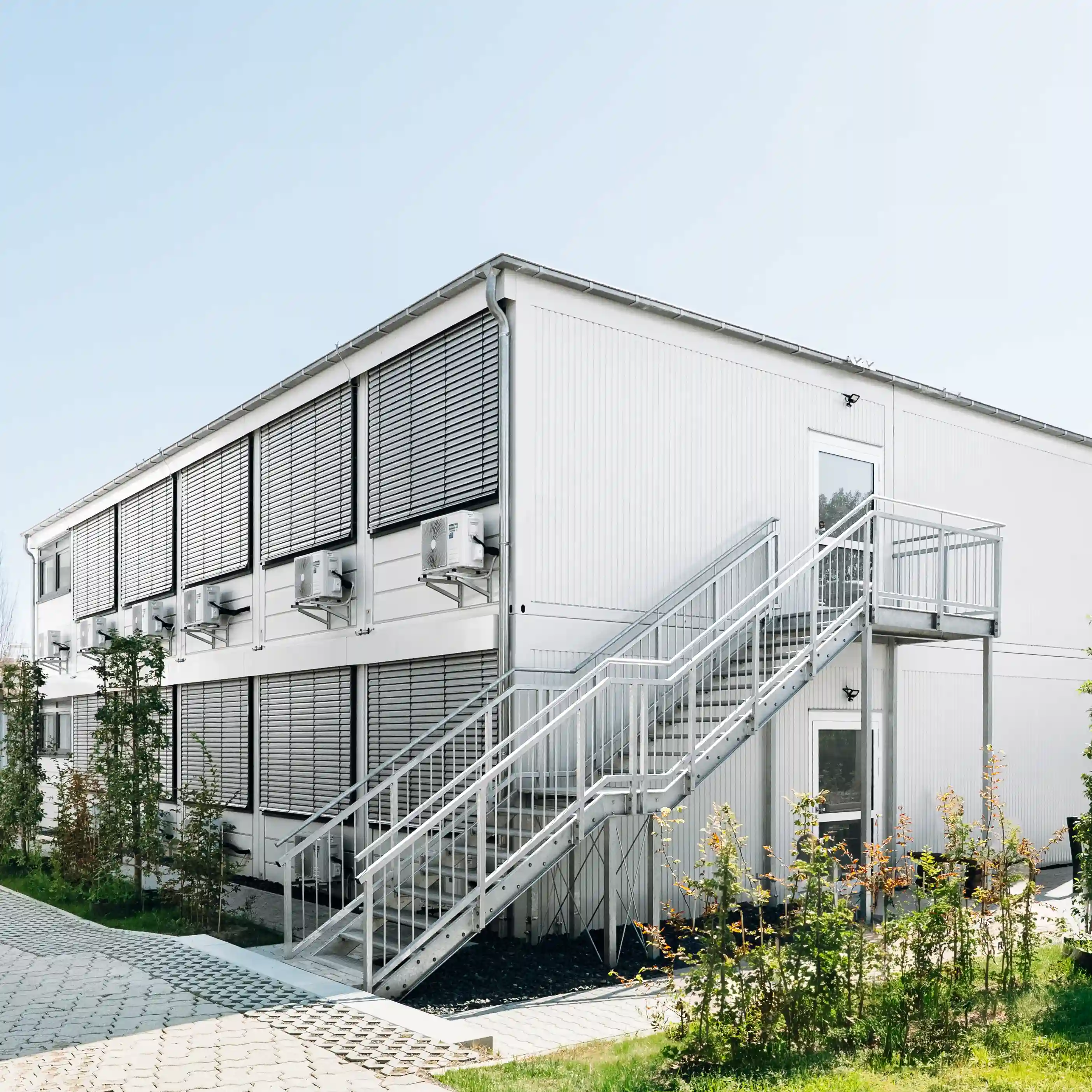 Zweigeschossiges Containergebäude mit außenliegender Treppenanlage, Sonnenschutzsystemen und moderner Gebäudetechnik. Das Gebäude ist von Grünflächen und befestigten Wegen umgeben.