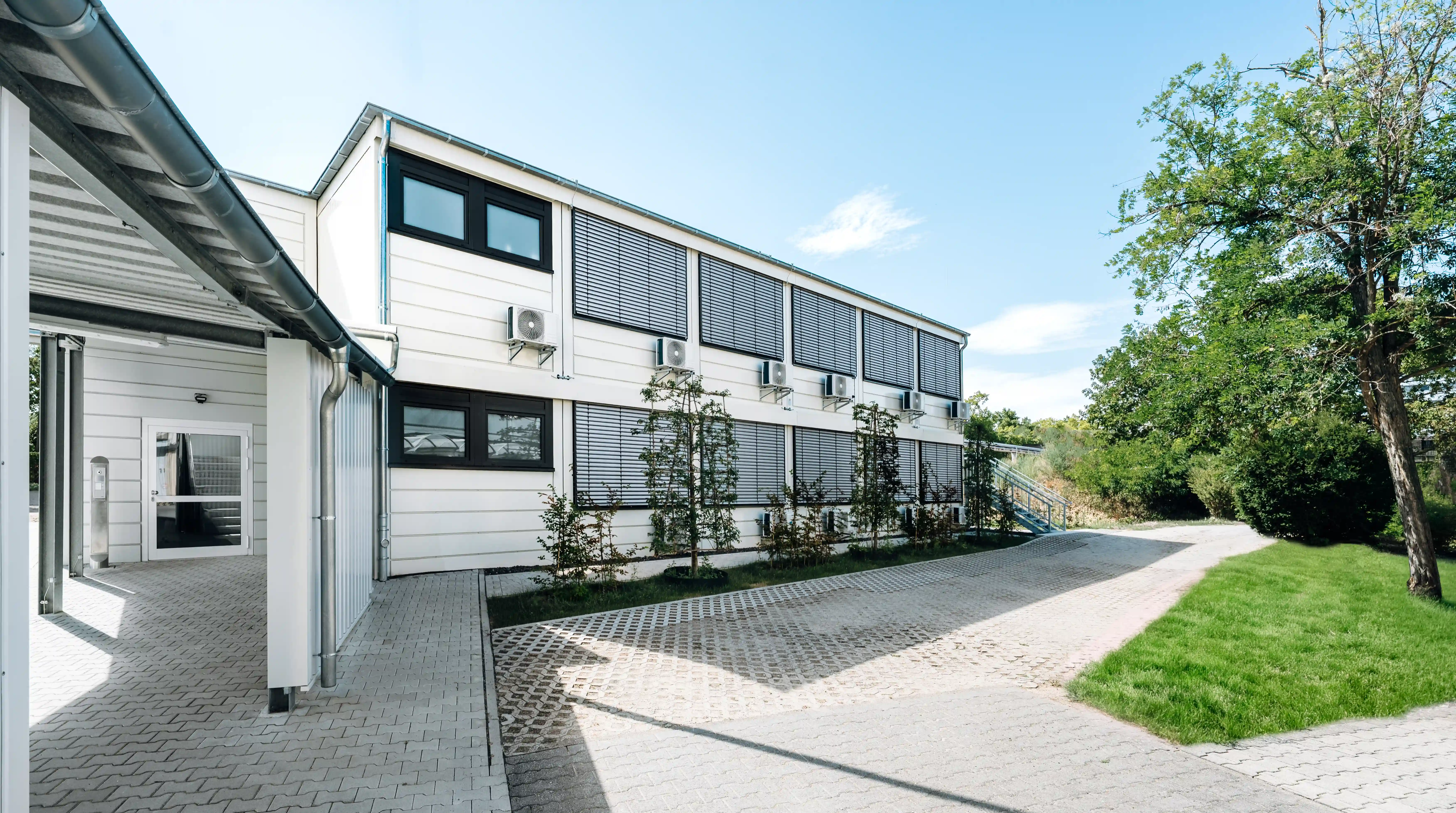 Zweigeschossiges Modulgebäude mit Sonnenschutzanlagen und Klimageräten an der Fassade. Das Gebäude ist von befestigten Wegen, Grünflächen und Bäumen umgeben.