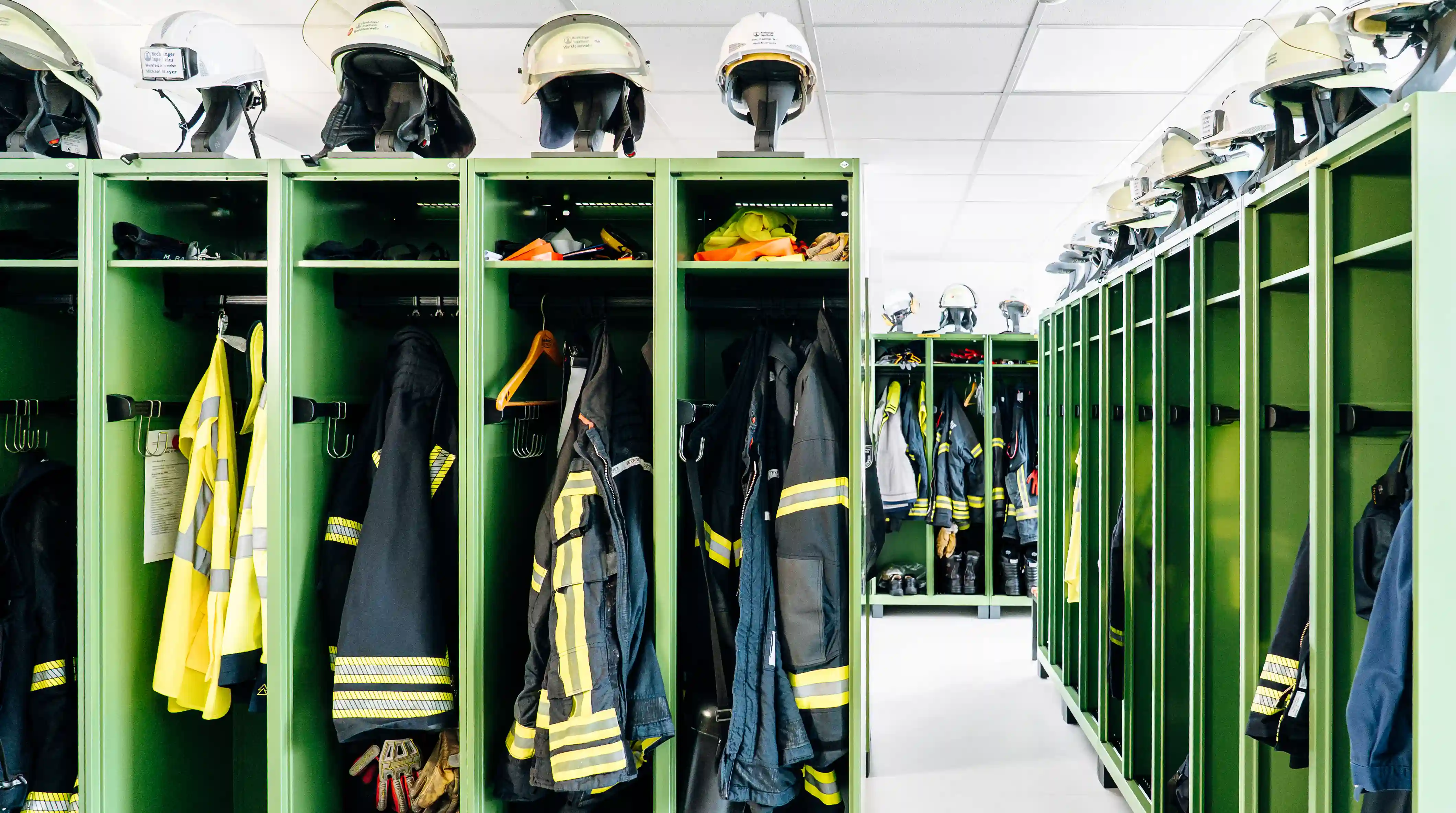 Umkleideraum einer Werk- oder Betriebsfeuerwehr mit grünen Spinden, Einsatzkleidung und Feuerwehrhelmen. Die Ausstattung ermöglicht die geordnete Aufbewahrung persönlicher Schutzausrüstung.