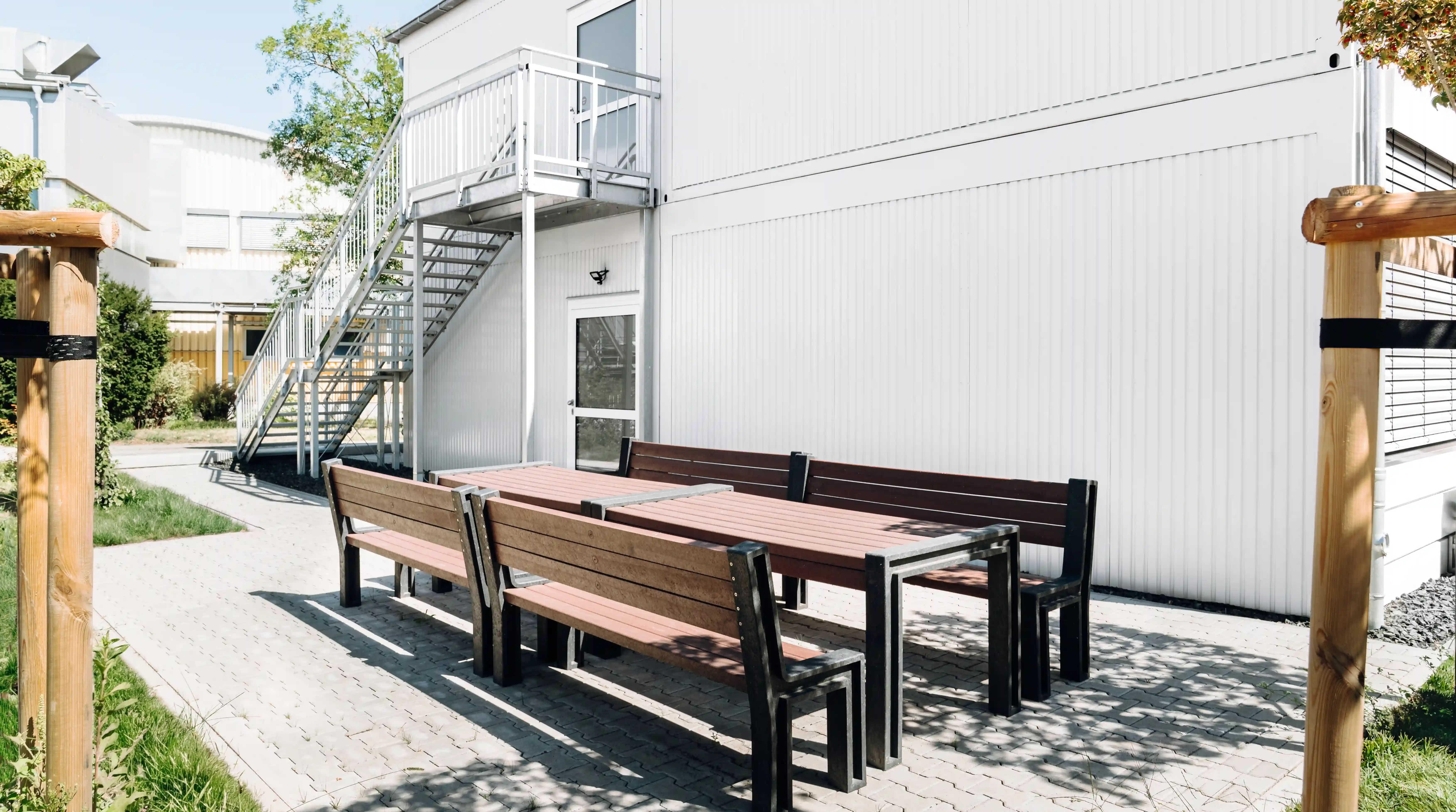 Außenbereich eines modularen Gebäudes mit Sitzgruppe aus Holzbänken und Tischen auf gepflasterter Fläche. Eine Außentreppe führt zu einem oberen Gebäudebereich.