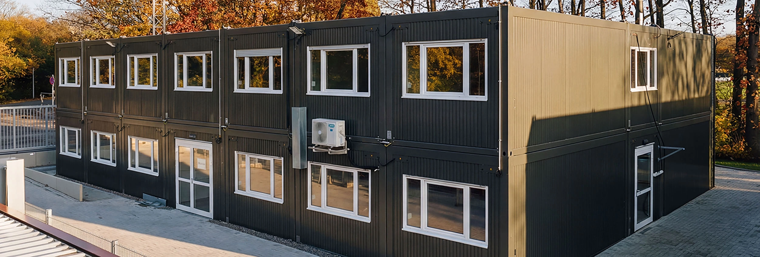 Zweigeschossige schwarze Containeranlage mit vielen Fenstern vor herbstlichen Bäumen