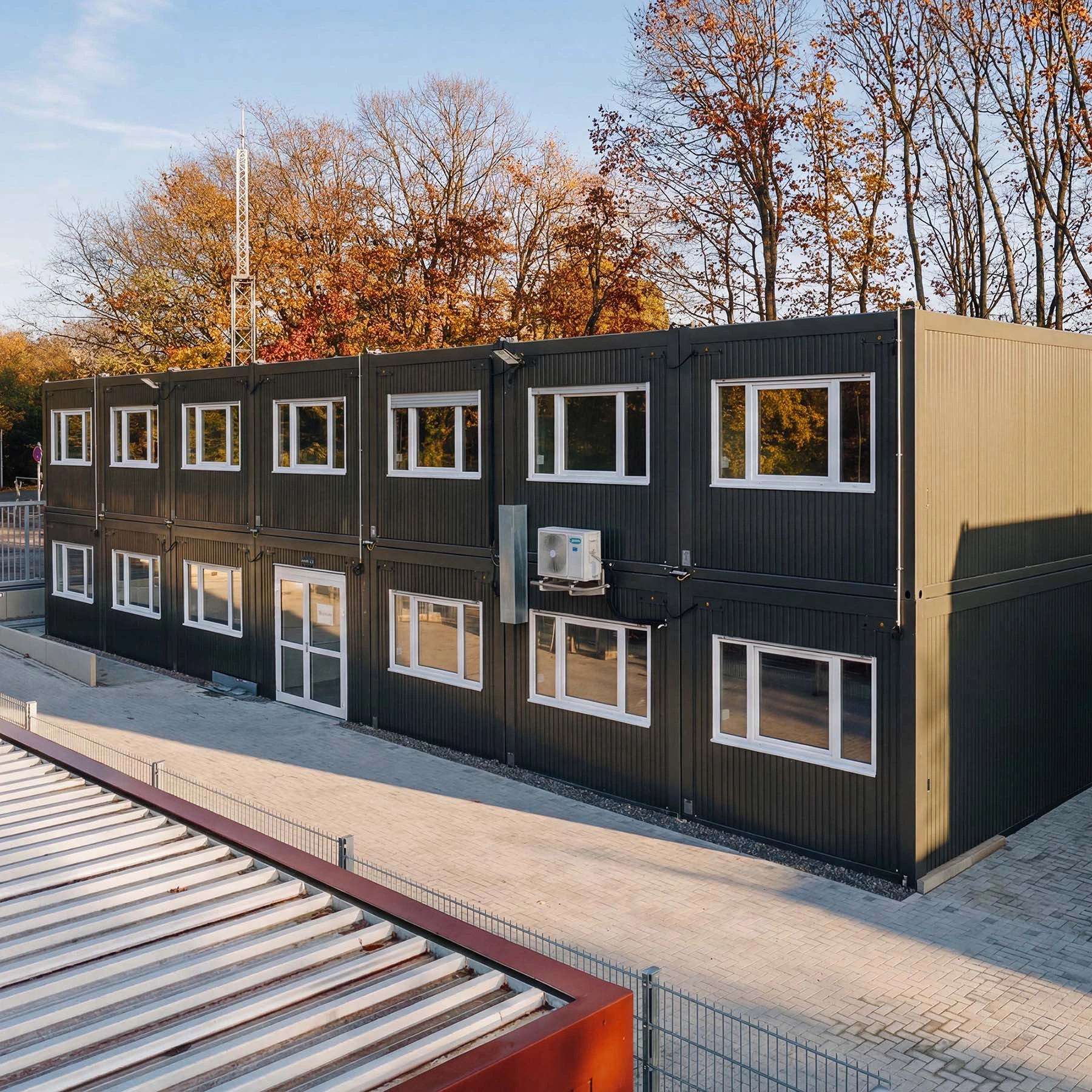 Zweigeschossige schwarze Containeranlage mit vielen Fenstern vor herbstlichen Bäumen