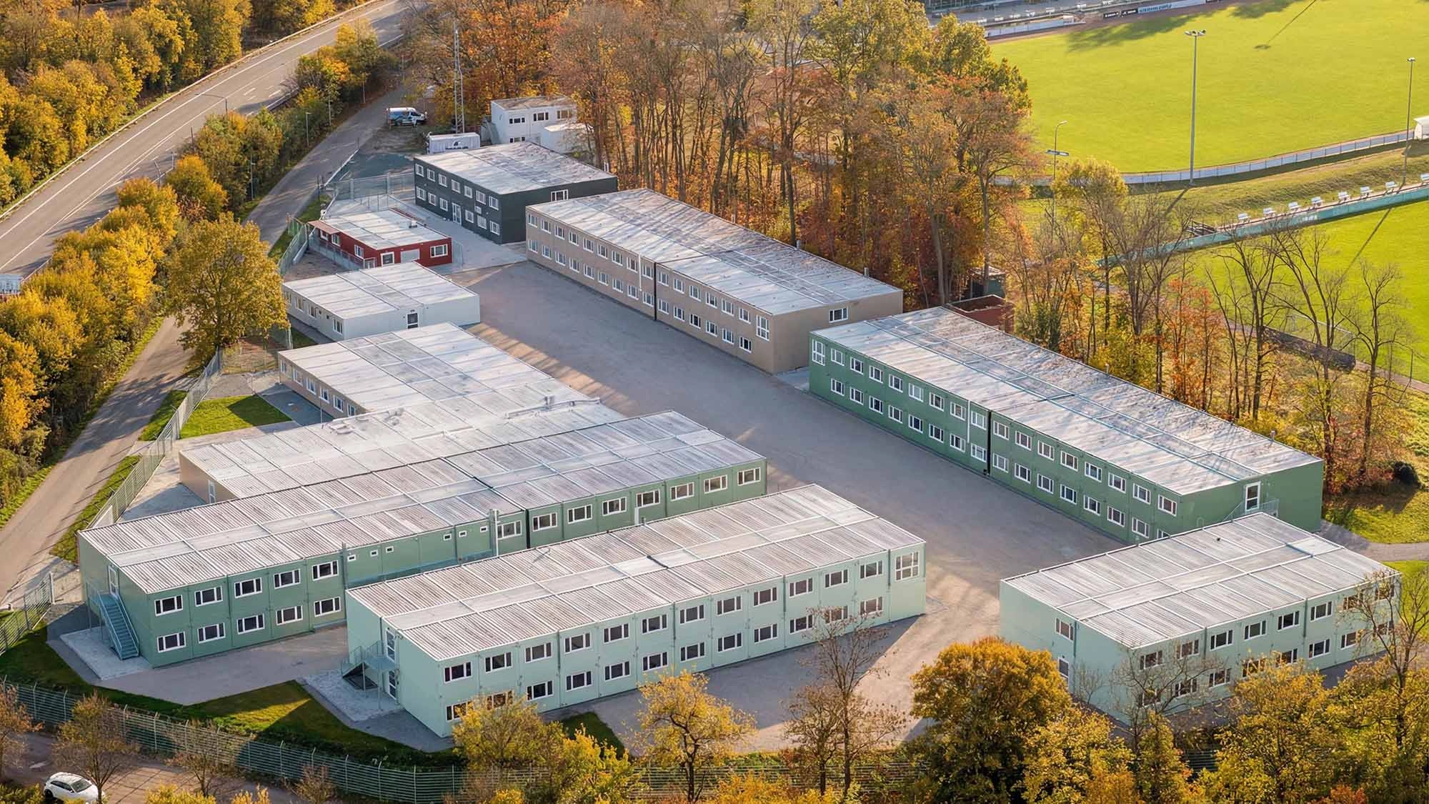 Luftaufnahme einer großen Containeranlage mit mehreren Gebäudeteilen zwischen Straße, Wald und Sportplatz