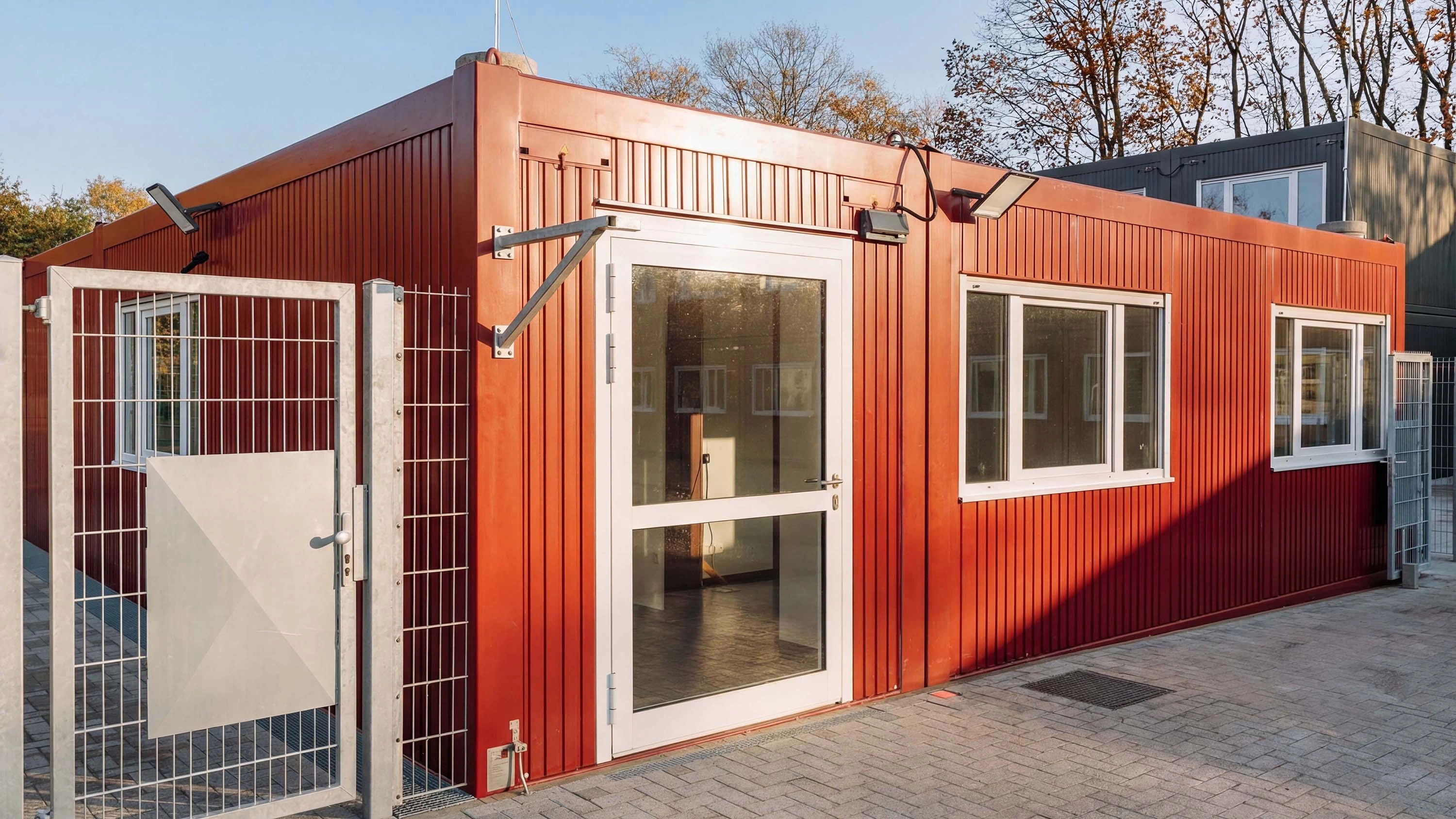 Rotes Containergebäude mit Glastür, Fenstern und umzäuntem Eingangsbereich