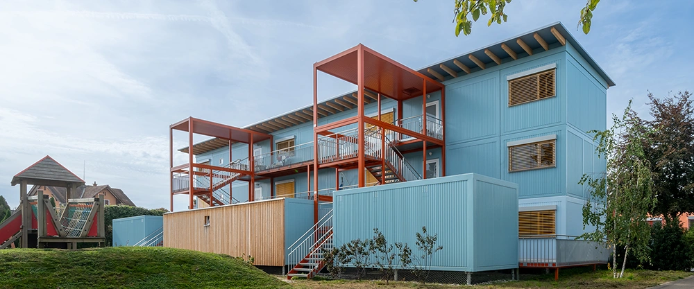 Ein blaues, dreistöckiges Modulgebäude zeigt orangefarbene Außentreppen und Balkone; es steht auf einer Rasenfläche neben einem Holz-Spielplatz in einer ruhigen Wohnumgebung unter klarem Himmel.