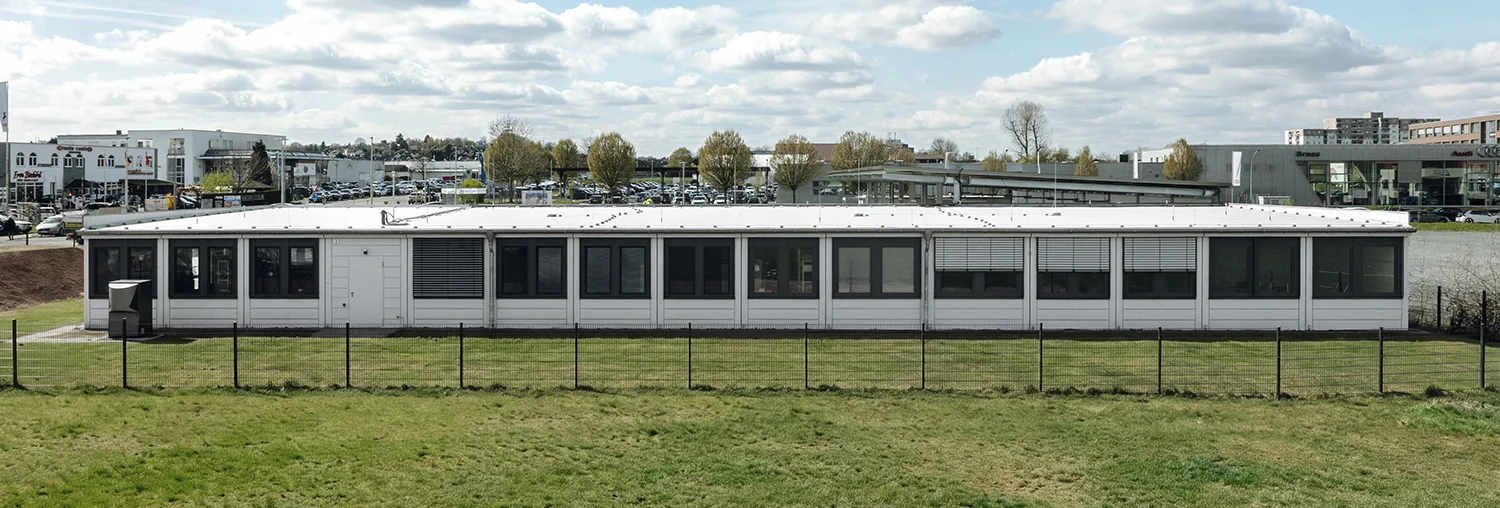 Eingeschossiges Modulgebäude mit weißer Fassade und großen Fensterfronten, umgeben von Zaun und Wiese in städtischer Umgebung
